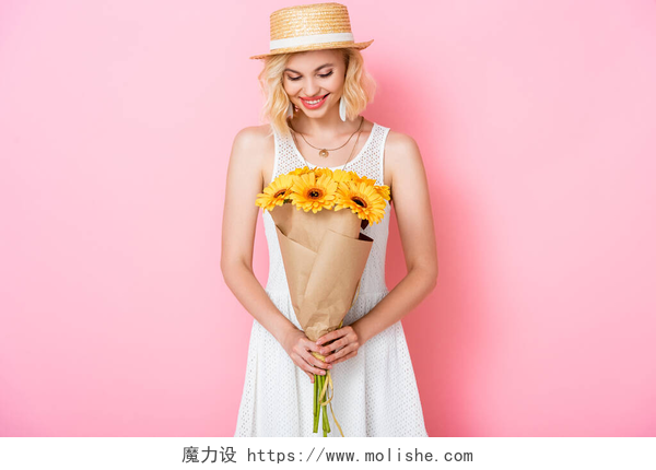 粉色背景上女孩手捧向日葵花束戴着草帽的年轻女子看着粉色的花朵 